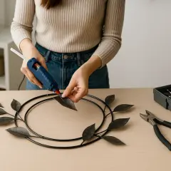 Kobieta przykleja liście do metalowej obręczy za pomocą pistoletu na klej. Obok leżą narzędzia.